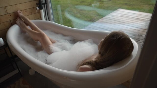 Young woman with long hair taking a relaxing bubble bath in a modern freestanding tub, enjoying the serene view of nature through a large window and finding tranquility at home
