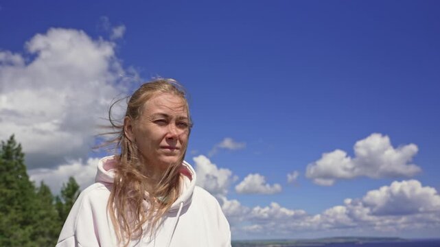 Thoughtful middle-aged woman with blonde hair blowing in the wind enjoying a beautiful sunny day with a blue sky and white clouds in the background, showing a range of emotions