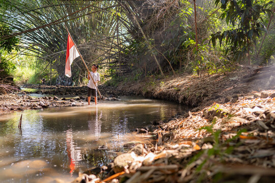 Young person stands in shallow woodland creek holding red and white flag beside leaves, roots, and dense green canopy, with bright sunlight, reflections, and peaceful wilderness mood