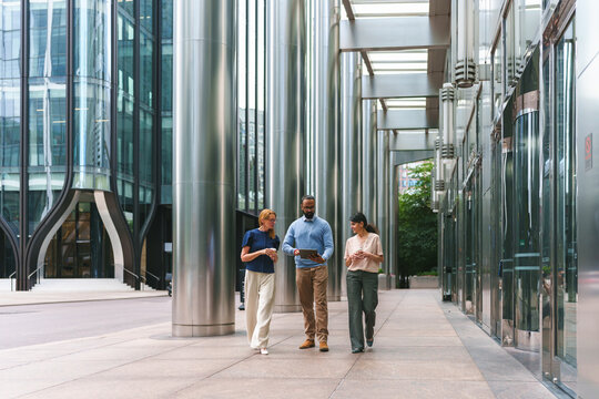 Diverse business professionals walking and discussing work while holding a tablet in a modern city setting