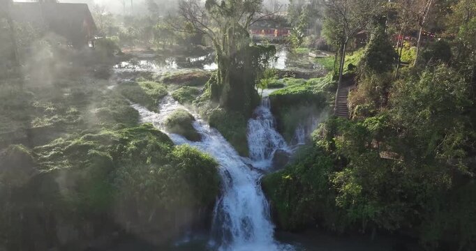 A dramatic drone zoom out shot revealing a magnificent waterfall cascading down into a lush, dense jungle with water splashing intensely and creating fine mist.