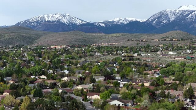 Suburban neighborhood spreads across high desert hills in Reno, Nevada, with tree-lined streets and houses beneath snow-capped Sierra Nevada mountains rising in distance. Aerial drone wide shot