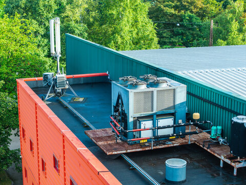 Aerial view of an industrial rooftop featuring a large HVAC chiller unit and a cell tower antenna, set against a backdrop of lush green trees in Riga, Latvia.