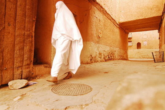 Algeria, Ghardaia, woman with white burka walking on narrow passageway with walls on both the side