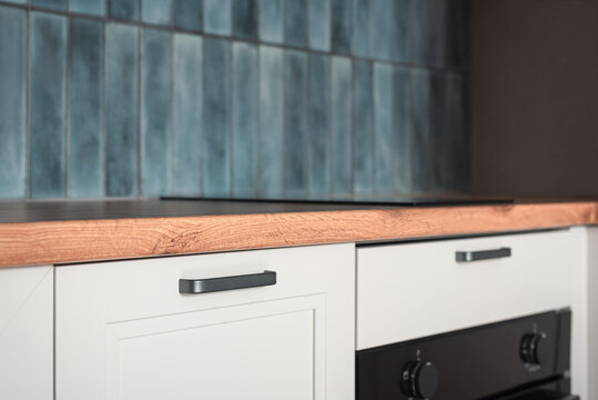 Close-up of a modern kitchen interior with wooden countertop, blue ceramic tile backsplash, white cabinets, black oven and induction cooktop. Contemporary home design.