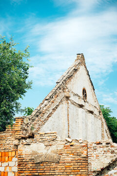 Weathered brick ruin building with a steep triangular gable and round window set against a bright blue sky, surrounded by trees and crumbling masonry that suggests age, history and decay.