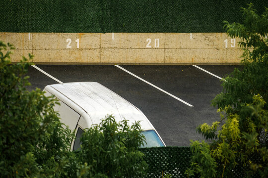 White delivery van parked in an empty parking lot during rainfall, viewed through green foliage. Urban transportation, logistics and rainy weather concept.