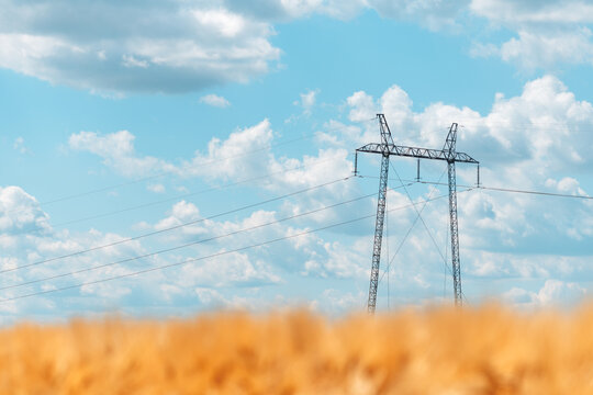 High-voltage transmission towers rise above a golden wheat field beneath a bright blue sky dotted with puffy white clouds, suggesting rural energy infrastructure