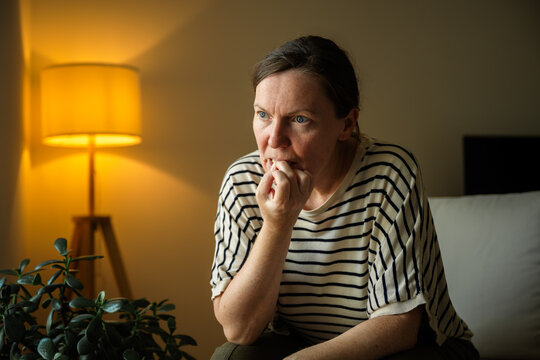 Middle-aged woman nervously biting her nails while sitting at home in the evening. Anxiety, stress, uncertainty and emotional distress concept.