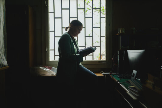 Silhouette of a woman standing by window in old library office reading a book, soft daylight and quiet academic atmosphere