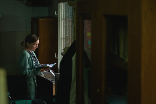 Female figure in low-lit old office room holding open book by the window, calm reading scene with moody atmosphere