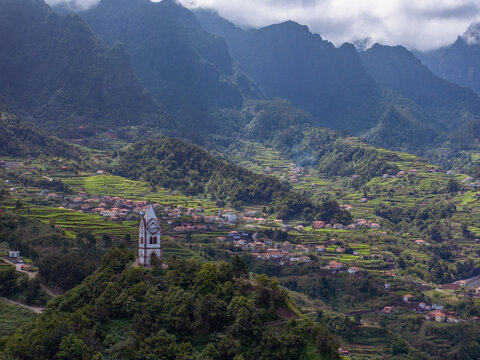 Aerial view of a historic clock tower standing on a lush green hill surrounded by terraced agricultural fields and small houses nestled in the mountainous landscape of Madeira.
