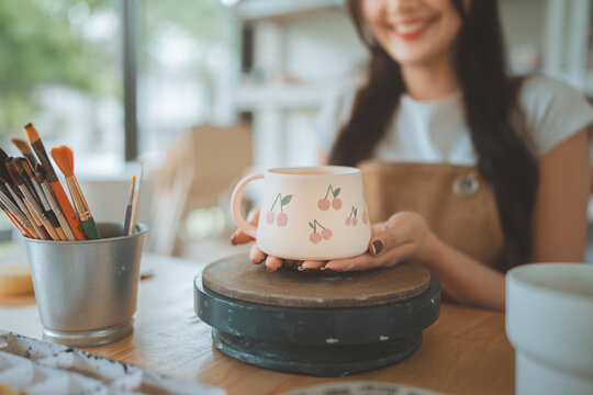 Close up of smiling Asian woman hands showing cute handmade ceramic mug with cherry pattern at pottery workshop, female artisan crafting clay cup for creative hobby, small business entrepreneur.
