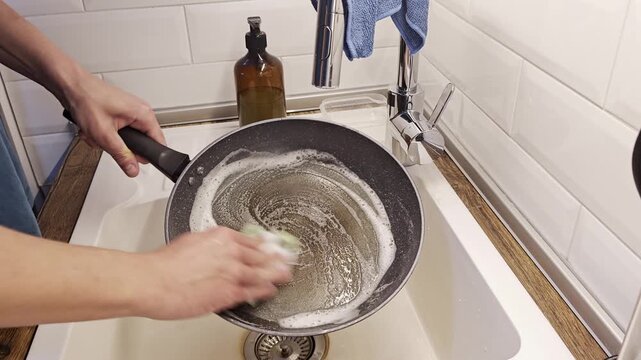Close up of a man scrubbing a black non-stick frying pan with a soapy sponge in a bright modern kitchen sink with dish soap bottle visible in background