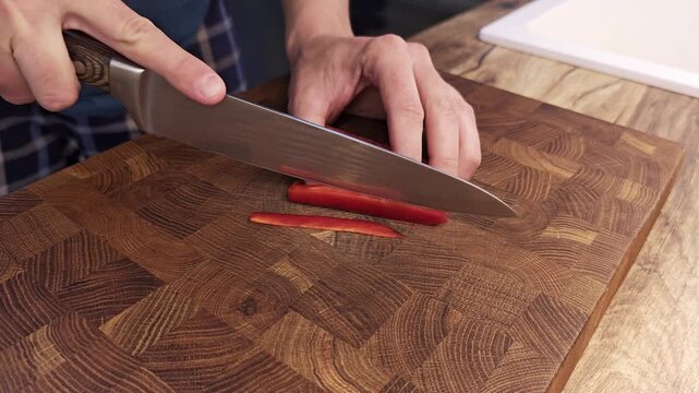 Close up of a man slicing fresh red bell peppers with a sharp knife on a wooden cutting board in a bright clean modern home kitchen