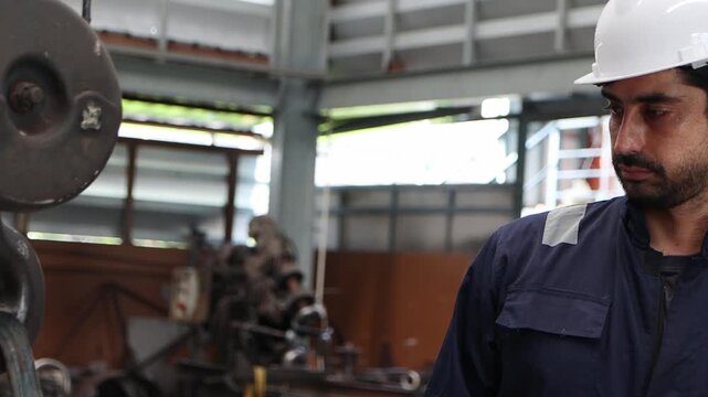 Industrial engineer supervisor foreman in safety suit and white helmet working to control machinery to lift a large iron hook up at manufacturing plant. Worker operates a lift machine in metal factory
