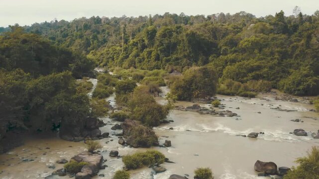 A stunning high-angle drone shot capturing a scenic waterfall flowing through a lush green jungle, flanked by dense rows of forest trees, featuring several large rocks covered in greenery protruding h