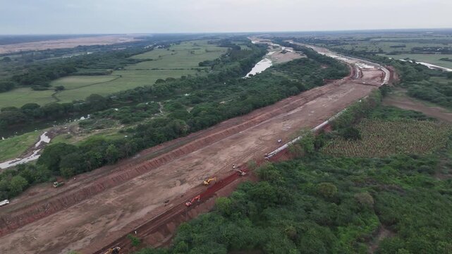 Wide aerial drone shot of the Grand Anicut Canal reconstruction work stretching across the frame. Freshly graded red laterite earth lines the long canal corridor with yellow excavators machinery.