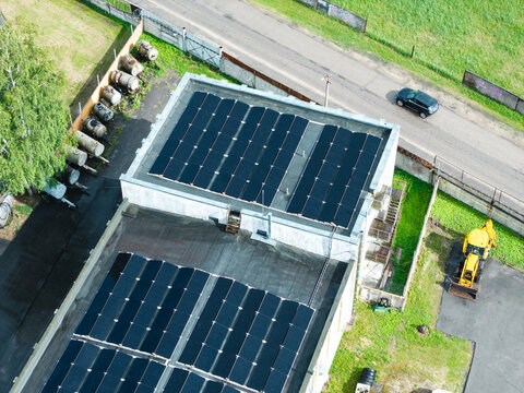Aerial view of solar panels installed on a flat industrial building roof with a yellow backhoe loader parked nearby and storage tanks in Olaine, Olaine Municipality, Latvia.