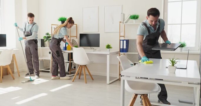 Team janitors cleaners mopping wiping office desks. Uniformed staff sanitize workstations, dust electronics and organize supplies in a workspace. Concept of professional facility maintenance.
