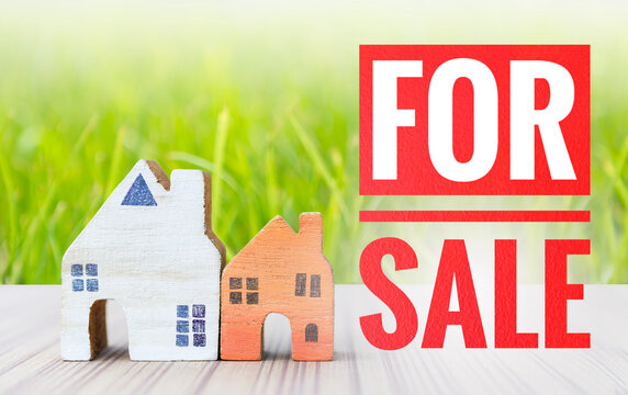 For sale sign with wooden house model over blurred green grass background, property and real estate business concept