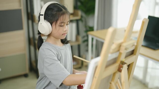 Young girl listening to music with headphones and painting on an easel in a bright art studio or classroom setting