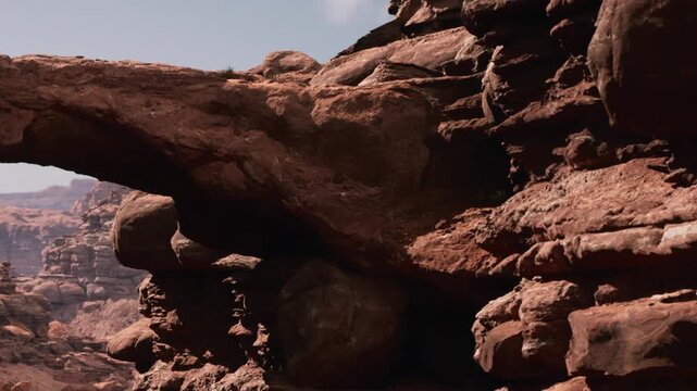 A large rock arch stands tall amidst a rugged desert landscape. The sun shines brightly, casting shadows on the rocky ground. The scene is filled with various shapes of rocks and formations.