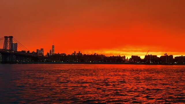 Williamsburg Bridge and Lower Manhattan skyline across East River. Steel bridge spans dark water with illuminated buildings below a red cloud layer.