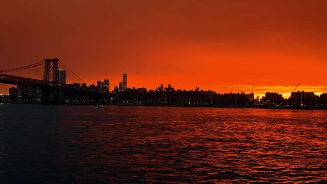 Williamsburg Bridge frames Lower Manhattan skyline across East River. Dark foreground water reflects city lights below a red cloud bank near dusk.