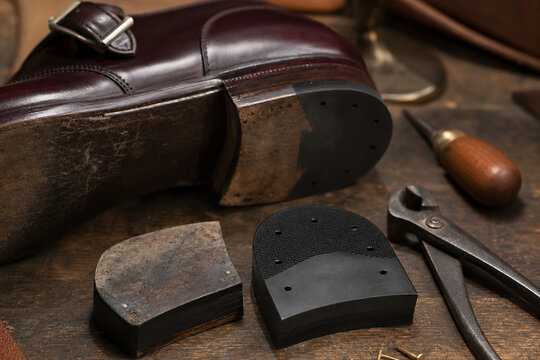 Close-up of a polished leather shoe, replacement soles, nails, and cobbler tools on a rustic wooden workbench in a traditional repair shop.
