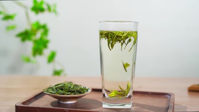 Fresh green tea leaves brewing in a tall glass cup on a wooden tray, with a small plate of dry tea leaves and a blurred green plant in the background.