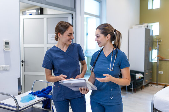 Two female doctors or surgeons in blue scrubs reviewing medical records in a hospital corridor. They are professional colleagues having a positive discussion about patient care 