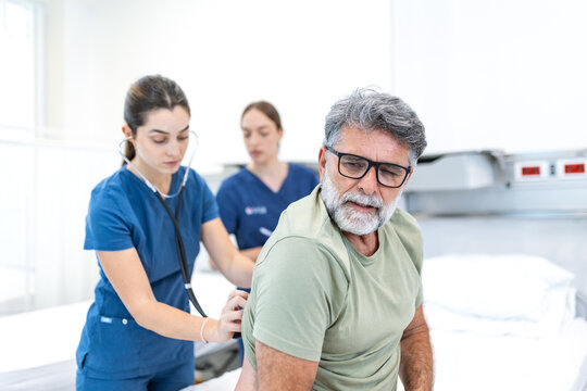 Female medical professional in blue scrubs using a stethoscope to examine the back of an older male patient in a clinical room. A colleague stands behind, observing and recording patient health data.