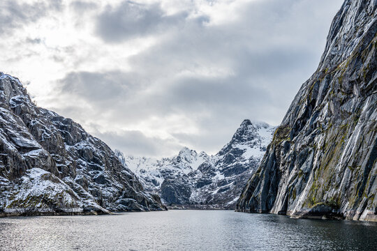 Trollfjord in Norway shows steep cliffs and snowy peaks surrounding the water on a cloudy day with reflections on calm water