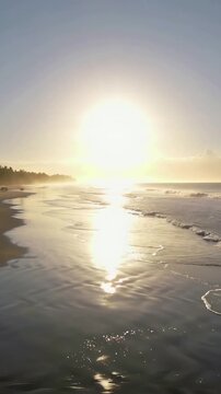 Vertical video: Shining sun nearing horizon casting long reflection on wet sand at beach, with surf