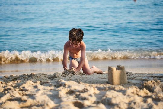 A focused child kneels on the sandy shore, shaping wet sand near gentle breaking waves during a family summer vacation.