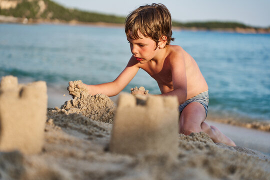 A focused child constructs sandcastles on a sandy shore next to the calm blue ocean during a family holiday.