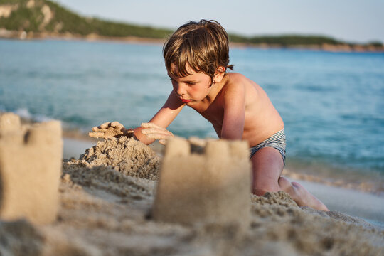 A focused child constructs sandcastles on a sandy shore next to the calm blue ocean during a family holiday.