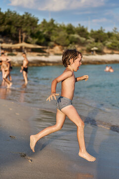 An energetic young kid sprints joyfully along the shoreline of a sunny sea resort, splashing near the water.