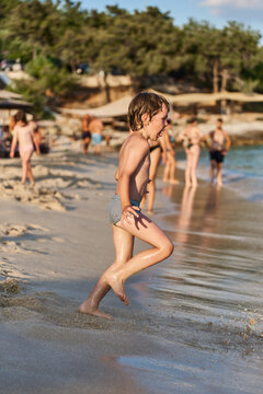 An energetic young kid sprints joyfully along the shoreline of a sunny sea resort, splashing near the water.