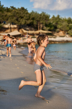 An energetic young kid sprints joyfully along the shoreline of a sunny sea resort, splashing near the water.
