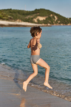An energetic young kid sprints joyfully along the shoreline of a sunny sea resort, splashing near the water.