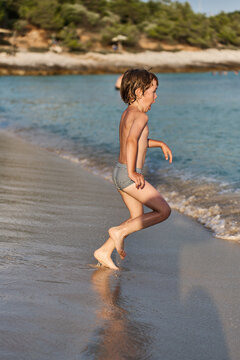 An energetic young kid sprints joyfully along the shoreline of a sunny sea resort, splashing near the water.
