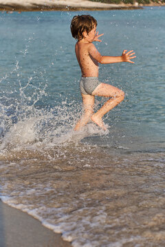 A cheerful child enjoys his summer vacation by sprinting through the shallow ocean waves, creating a dynamic splash of water.
