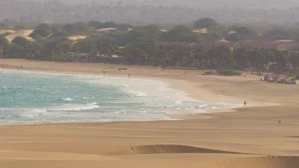 a vast coastal landscape under a hazy, overcast sky. In the foreground, smooth, rolling sand dunes with soft ridges stretch across the scene, transitioning into a wide, sandy beach.  © kapros76