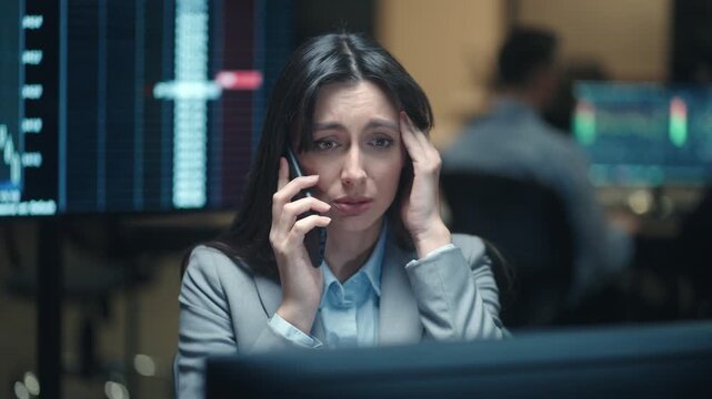 Concerned Caucasian female broker talking on smartphone and touching forehead near financial charts. Woman trader reacting to stock market loss in dark office. Stressful work call at night.