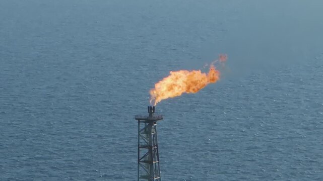 Close drone footage of a burning flare tower on an offshore oil production platform set against deep ocean water