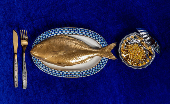 A surreal golden fish and caviar sit on a blue velvet table