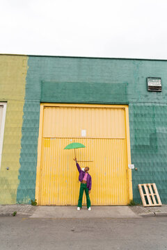 Non-binary person holding green umbrella in front of yellow shutter door