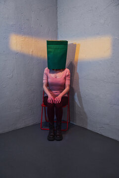 Woman hides her face inside a green paper bag in a corner